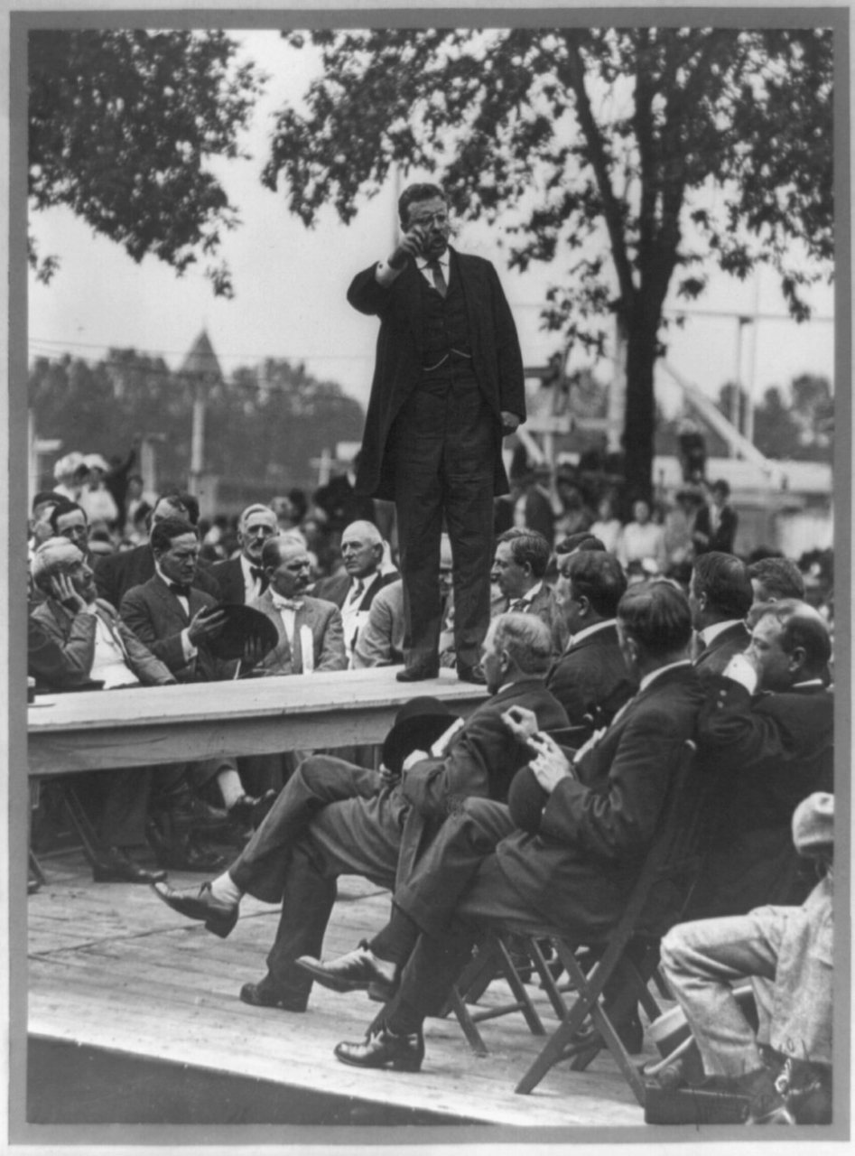 Theodore Roosevelt standing on table outdoors and making speech to men seated around him, right hand raised) - Smith Ar…