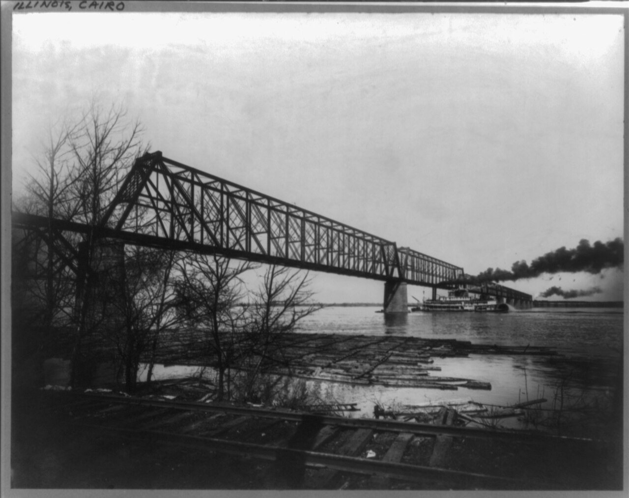 Stern-wheel riverboat passing under Illinois Central Railroad bridge over Ohio River, Cairo, Illinois, with logs lashed…