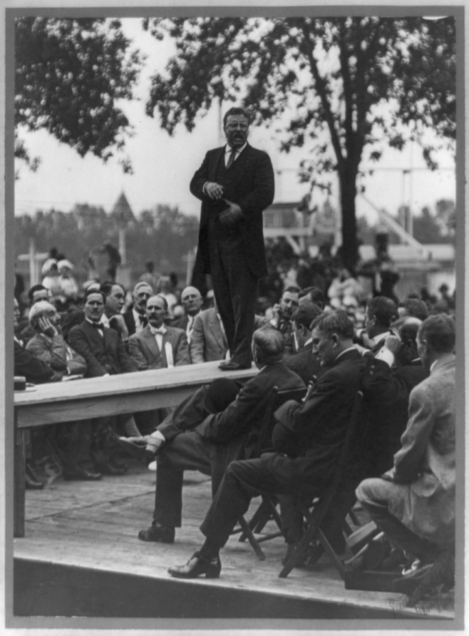 Theodore Roosevelt standing on table outdoors and making speech to men seated around him, hands at waist) - Smith Art P…