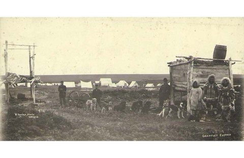 Dogsled team hauling cart, possibly the Grantley Express, with Eskimo man and two Eskimo women at right, Teller, Alaska…